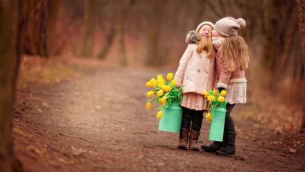 Two smiling children share a joyful kiss while holding buckets of yellow flowers, set against a serene pathway in a warm, autumnal landscape. A heartwarming photography background.