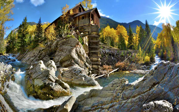 HD desktop wallpaper showcasing the man-made Crystal Mill perched on rocky terrain beside a flowing river, surrounded by autumn trees and a bright sunburst in a mountain landscape.