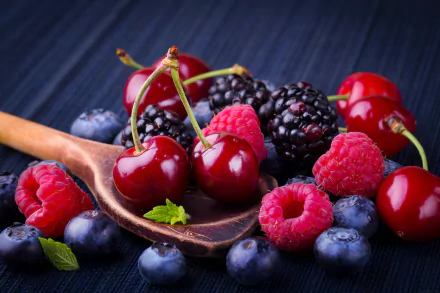 A vibrant HD desktop wallpaper featuring fresh cherries, blackberries, blueberries, and raspberries arranged on a dark background with a wooden spoon. The image showcases a colorful assortment of berries and fruit.