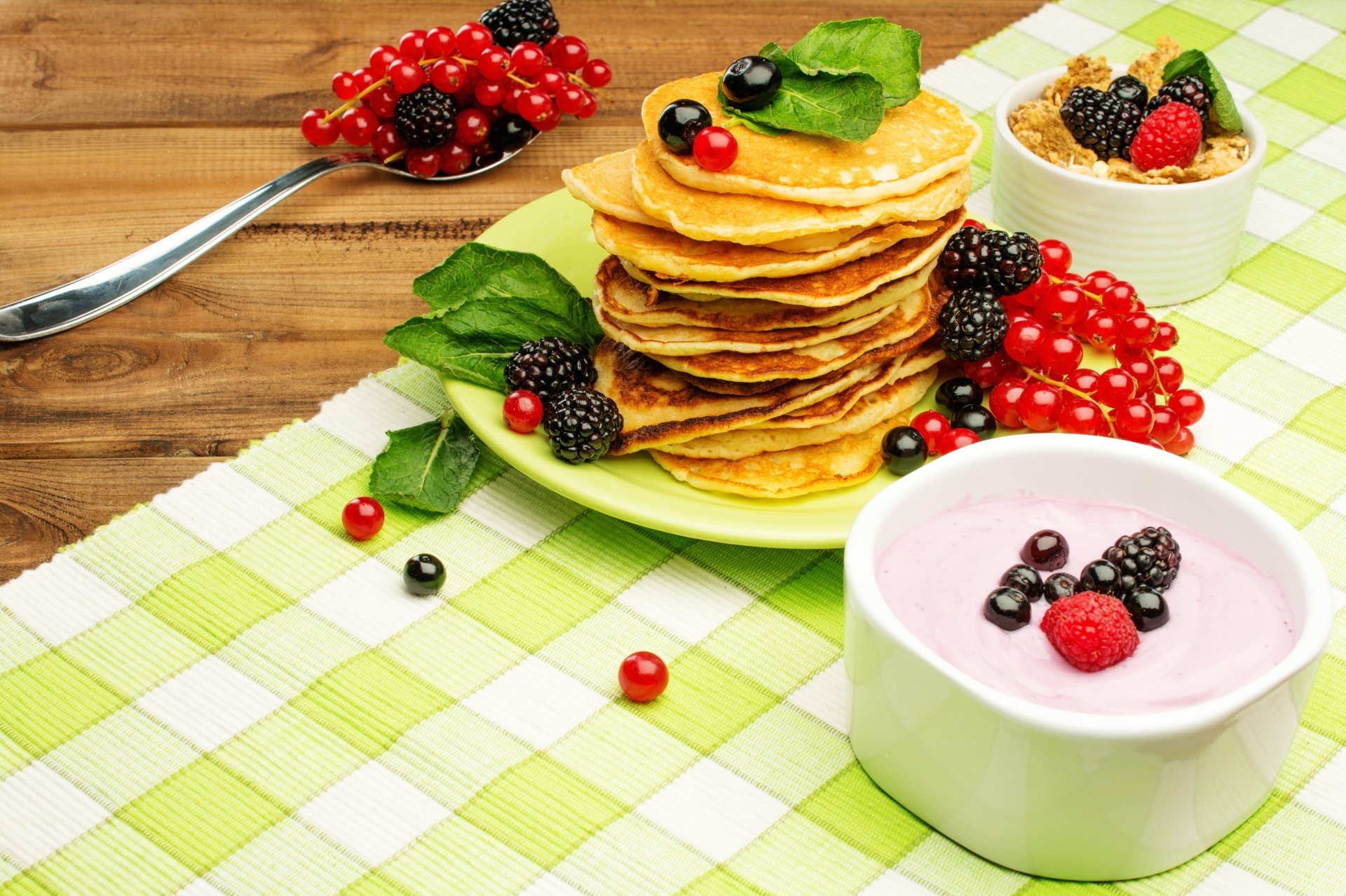 Stack of pancakes topped with currants, blueberries, raspberries, and blackberries, served alongside a bowl of berry yogurt and muesli on a checkered tablecloth.