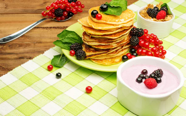 Stack of pancakes topped with currants, blueberries, raspberries, and blackberries, served alongside a bowl of berry yogurt and muesli on a checkered tablecloth.