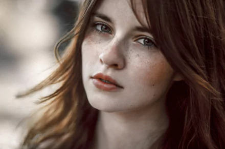 A brunette woman with freckles gazes directly at the camera, her flowing hair softly framing her face, creating an elegant and captivating HD desktop wallpaper.