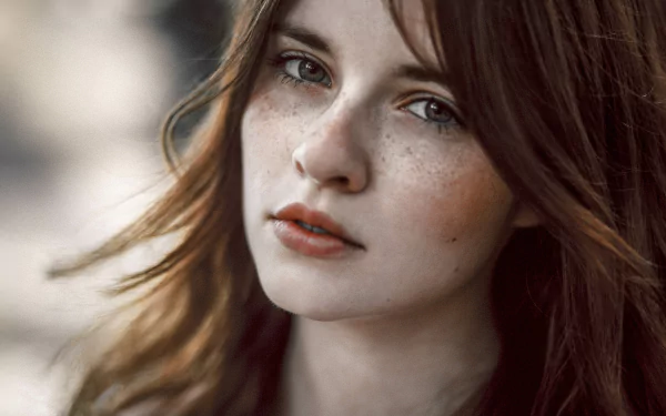 A brunette woman with freckles gazes directly at the camera, her flowing hair softly framing her face, creating an elegant and captivating HD desktop wallpaper.