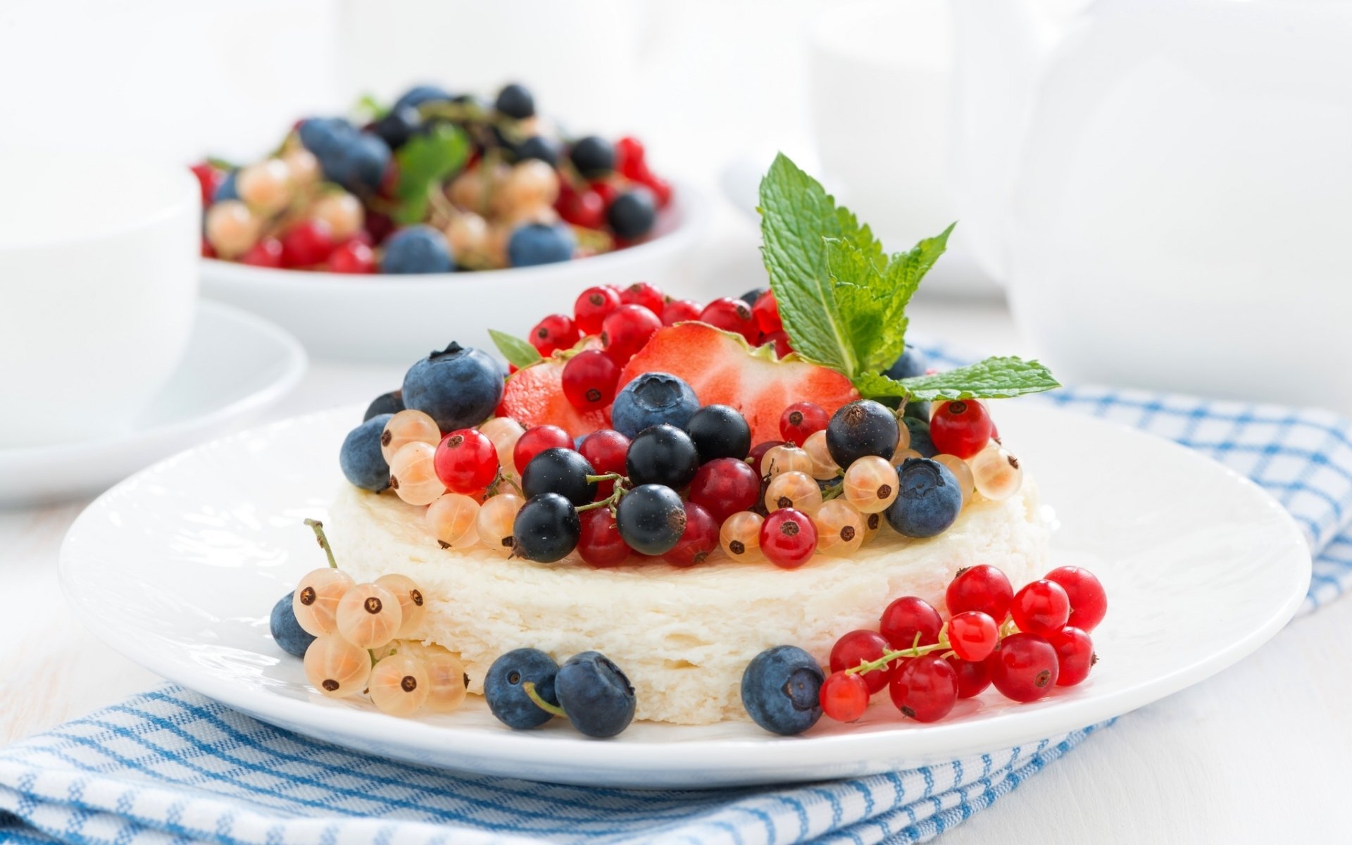 A vibrant cheesecake dessert topped with fresh currants, blueberries, and a mint leaf, set on a white plate with a blurred background.