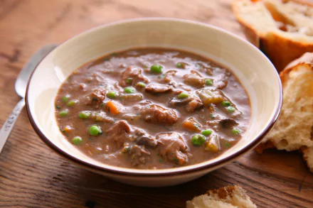 2K Quad HD PC desktop wallpaper: close-up of a hearty beef-and-vegetable stew in a bowl with peas and crusty bread on a wooden table.