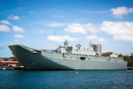 HMAS Canberra (L02), Royal Australian Navy amphibious assault ship and helicopter carrier, moored off a city shoreline under blue sky — large gray warship on calm water.