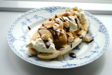 A close-up of a banana split topped with whipped cream, chocolate sauce, and sliced nuts on a decorative plate, captured in 4K Ultra HD quality.