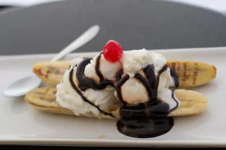 HD desktop wallpaper of a banana split dessert featuring ice cream, whipped cream, chocolate syrup, and a cherry on a white plate.