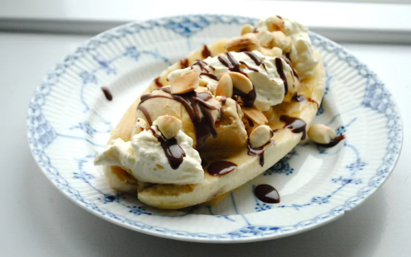 A close-up of a banana split topped with whipped cream, chocolate sauce, and sliced nuts on a decorative plate, captured in 4K Ultra HD quality.