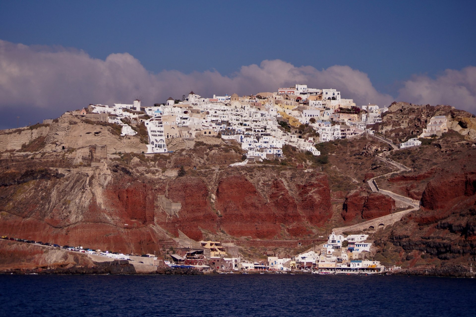 4K Ultra HD landscape of the man-made white buildings of Santorini island, Greece, perched on red cliffs above the blue sea under a partly cloudy sky.