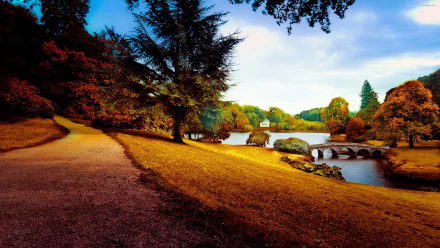 A vibrant park scene with a winding path, lush trees, and a serene lake, enhanced by autumn colors. Captured in stunning 4K Ultra HD for a striking desktop wallpaper.