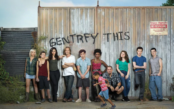 Group of diverse characters from the TV show Shameless pose in front of a weathered fence with the words Gentrify This spray-painted on it, capturing their unique dynamics and urban setting.