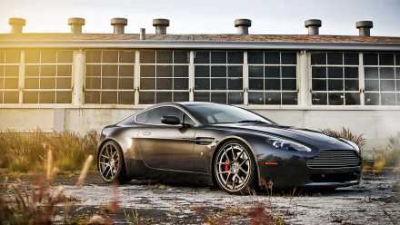 Sleek gray Aston Martin sports vehicle parked in front of an industrial building at sunset, 2K Quad HD PC desktop wallpaper and background.