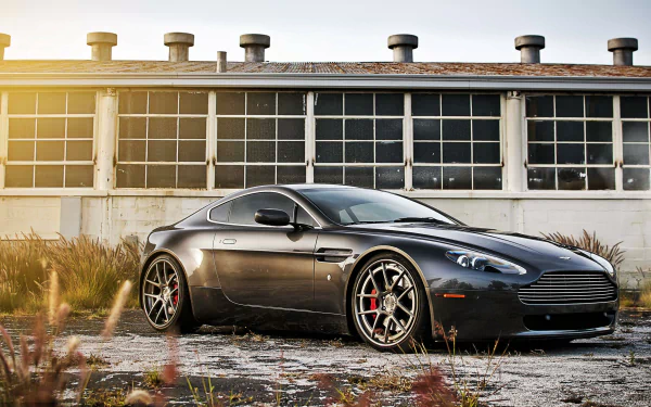 Sleek gray Aston Martin sports vehicle parked in front of an industrial building at sunset, 2K Quad HD PC desktop wallpaper and background.