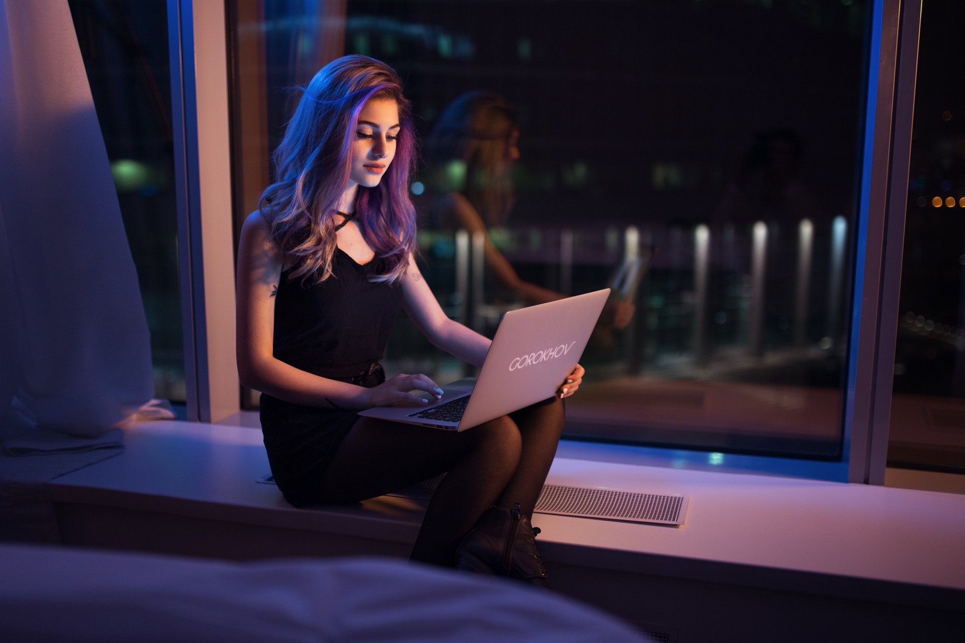 A woman with purple hair sits by a window in a dimly lit room, using a laptop. The cityscape outside adds a modern touch to this HD desktop wallpaper and background.