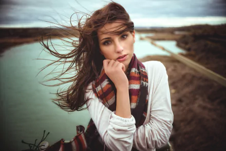 HD PC desktop wallpaper of a long-haired woman model with windblown hair and a striped scarf, posing thoughtfully outdoors by a river and distant bridge.