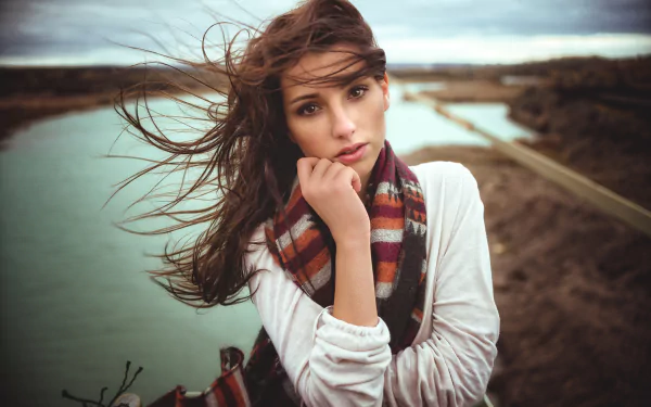 HD PC desktop wallpaper of a long-haired woman model with windblown hair and a striped scarf, posing thoughtfully outdoors by a river and distant bridge.