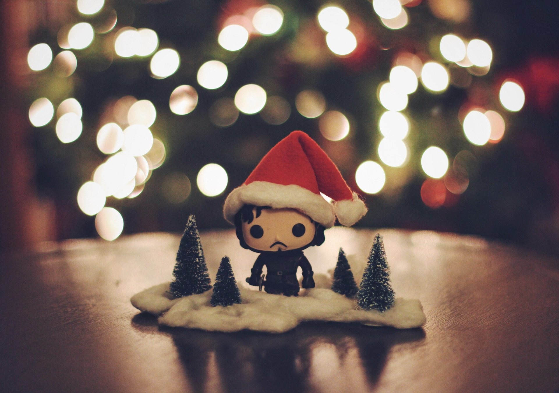 A festive Jon Snow toy, wearing a Santa hat, stands on snowy grass surrounded by miniature trees, with a bokeh background that evokes the enchanting world of Game of Thrones.