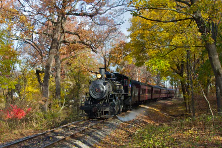 A classic steam train locomotive steaming through a colorful autumn landscape, surrounded by vibrant trees and foliage, creating a picturesque scene for a desktop wallpaper.