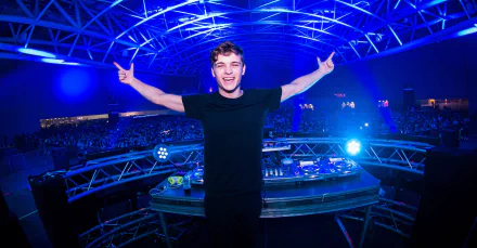 A vibrant HD wallpaper featuring DJ Martin Garrix performing energetically on stage, illuminated by striking blue lights and a cheering crowd in the background.