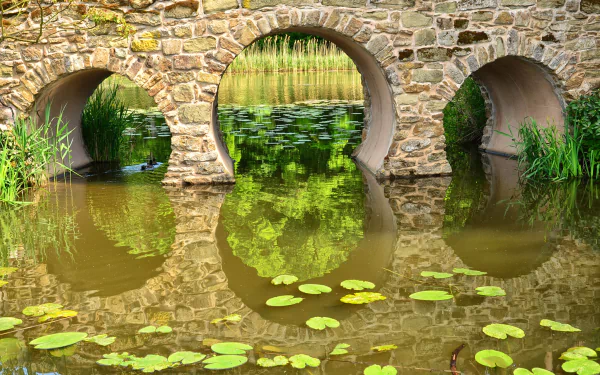 Stone arch bridge with three arches reflected in a pond surrounded by greenery, creating a serene, man-made landscape in this HD PC desktop wallpaper.