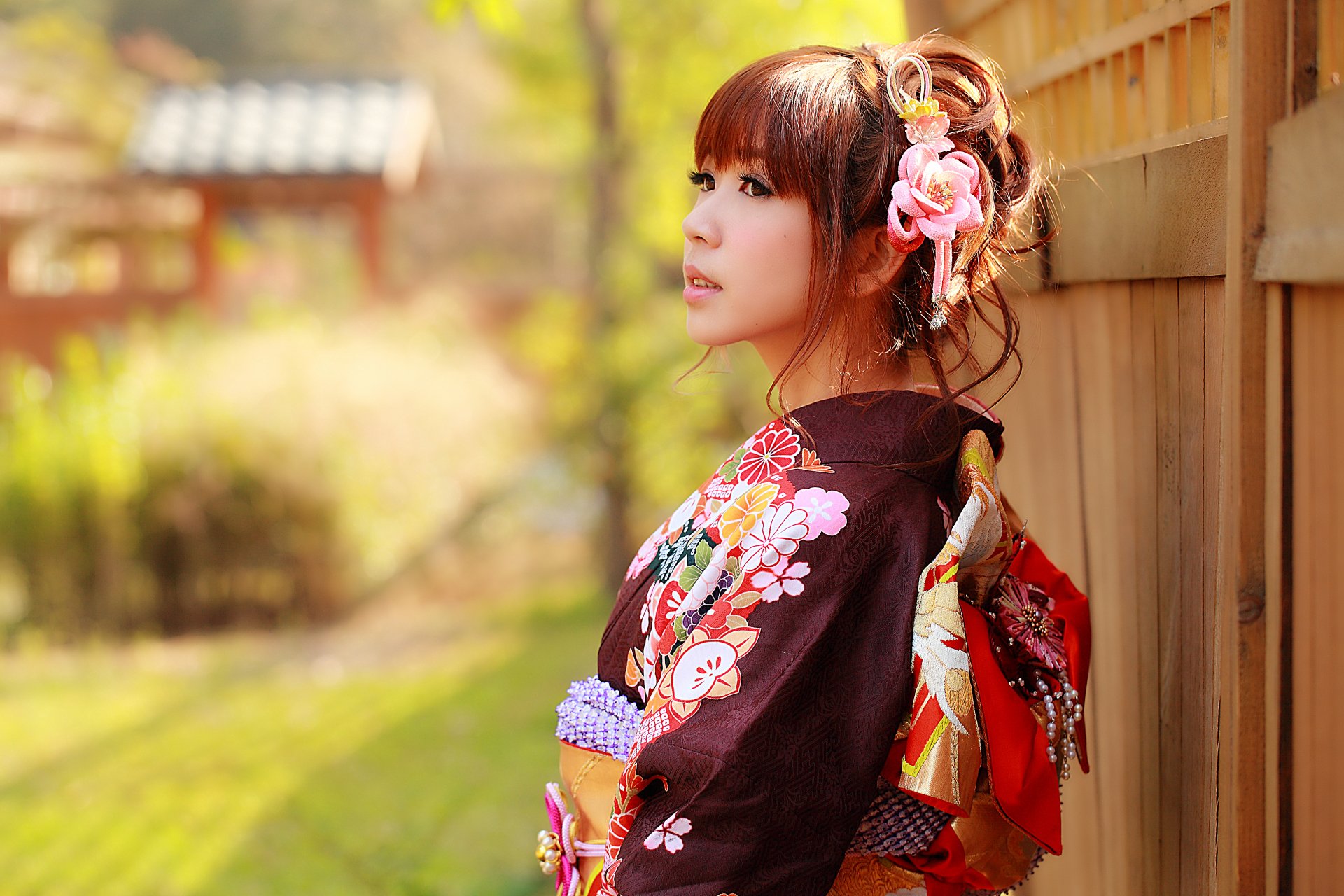 An Asian woman with brunette hair stands outside, wearing a colorful traditional costume with floral patterns. The background features blurred natural scenery and traditional architecture. HD desktop wallpaper.