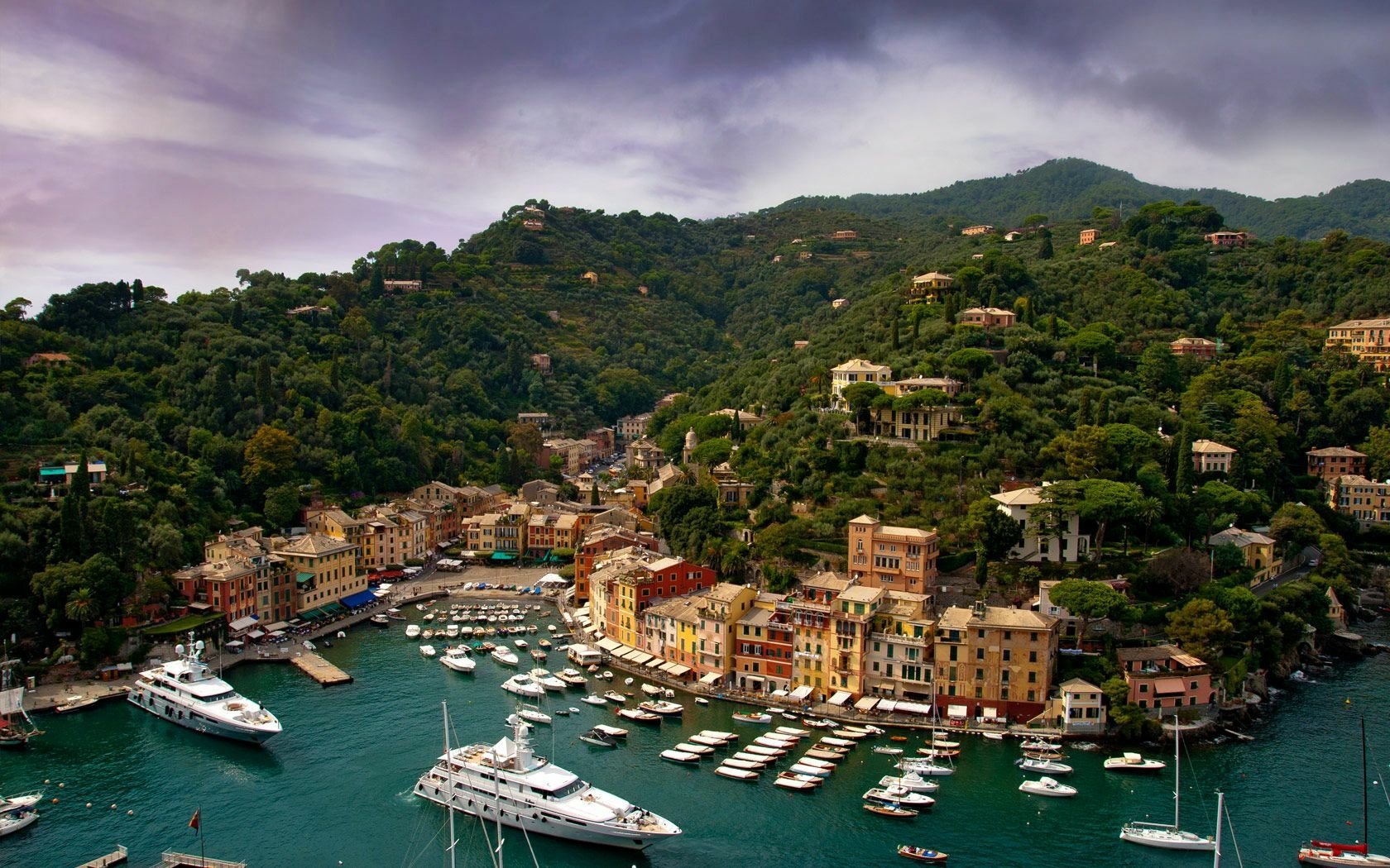 HD PC desktop wallpaper of Portofino harbor: colorful man-made waterfront buildings and yachts along the dock, framed by lush green hills beneath a dramatic cloudy sky.