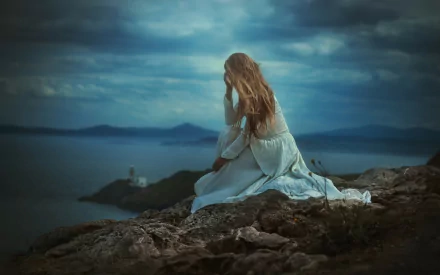 A blonde woman in a white dress sits alone on rocky coastline, exuding a sad and contemplative mood, against a scenic backdrop of dark, cloudy skies and distant sea horizon. HD desktop wallpaper.