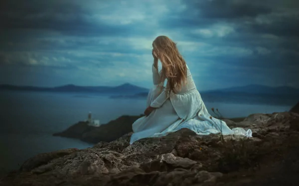 A blonde woman in a white dress sits alone on rocky coastline, exuding a sad and contemplative mood, against a scenic backdrop of dark, cloudy skies and distant sea horizon. HD desktop wallpaper.