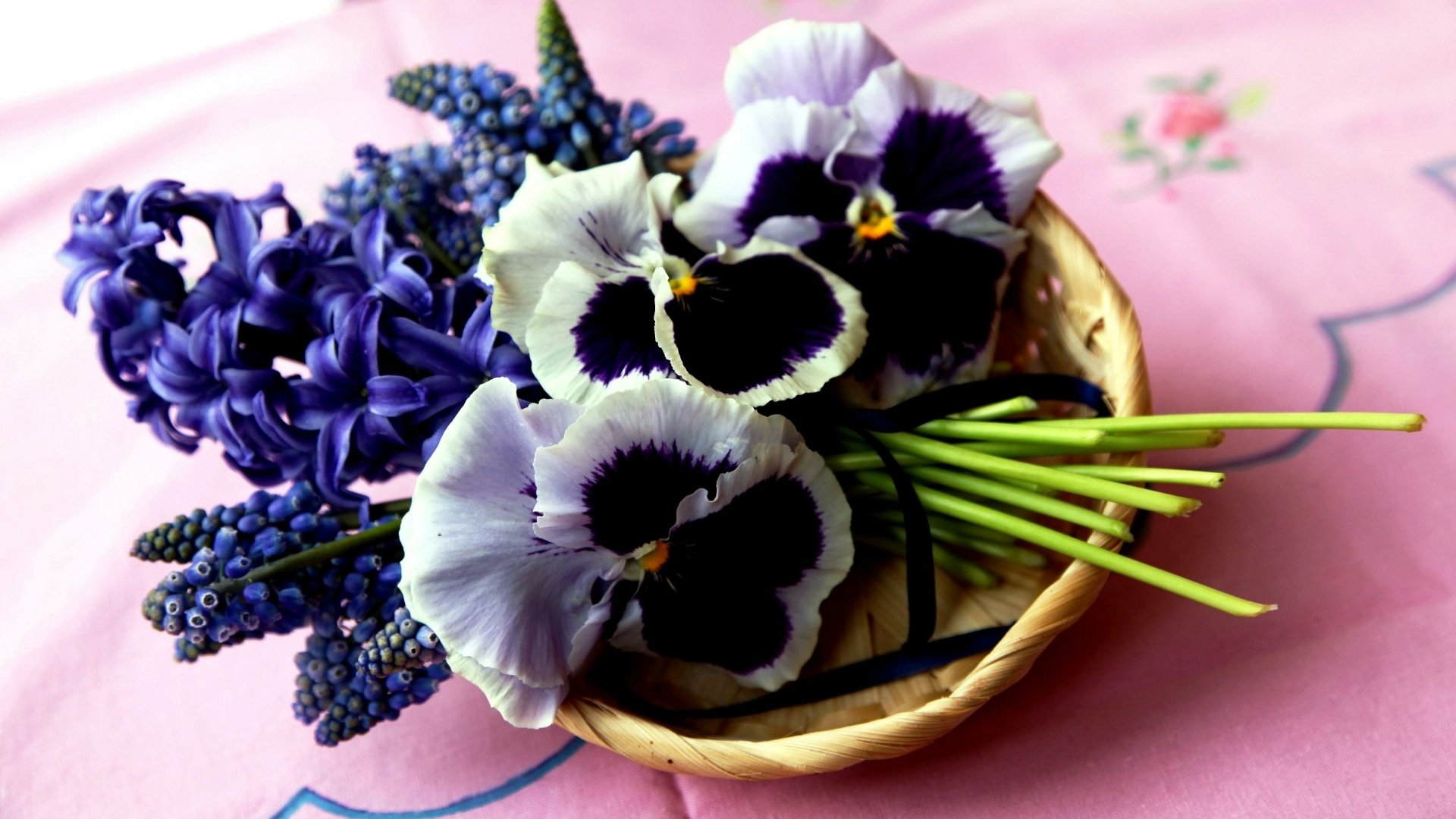 A vibrant 4K Ultra HD desktop wallpaper featuring a man-made arrangement of purple muscari, hyacinth, and pansy flowers in a woven basket on a pink floral cloth.