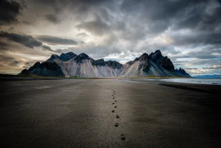 Footprints trail across a sandy beach toward rugged mountains under a dramatic cloudy sky, captured in an HD landscape wallpaper.