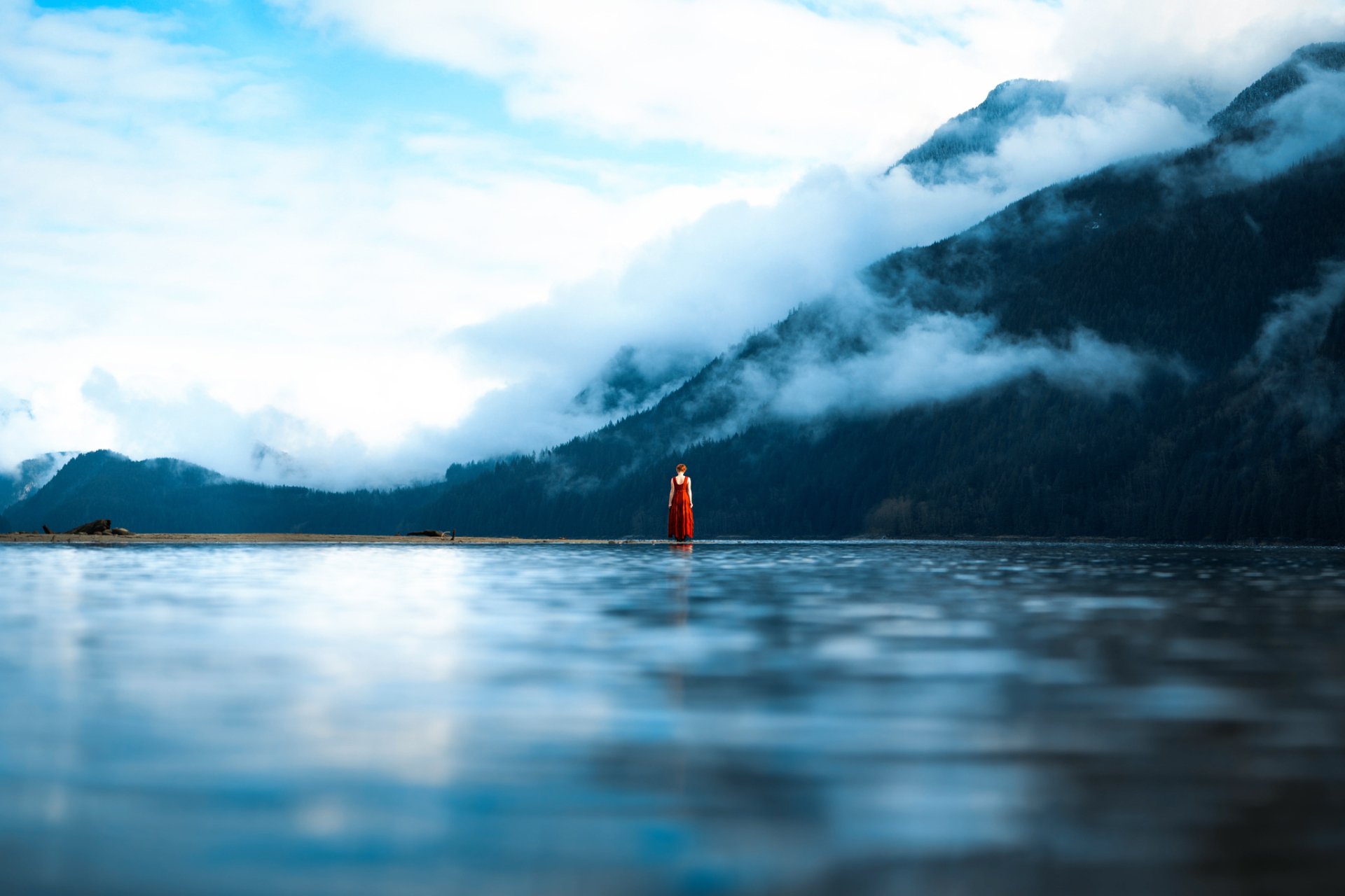 A woman stands with her back to the camera, facing a calm body of water with mountains in the background. Clouds drift down the slopes, creating a serene, HD scene for a desktop wallpaper.