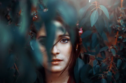 HD desktop wallpaper featuring a brunette woman with brown eyes partially hidden behind leaves, blending natural elements with a striking portrait.