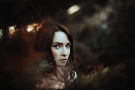 HD PC desktop wallpaper of a brunette model with brown eyes framed by soft bokeh and moody foliage, close-up portrait.