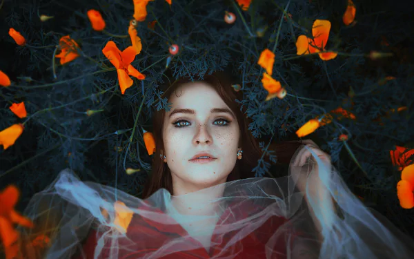 Brunette woman with freckles and brown eyes lies among vibrant orange flowers, wearing delicate earrings and a translucent veil in this HD desktop wallpaper.