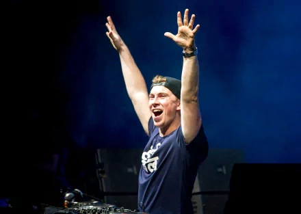 HD PC desktop wallpaper: energetic male DJ in a backward cap and navy tee, smiling with arms raised on a smoky blue concert stage — music-themed background.