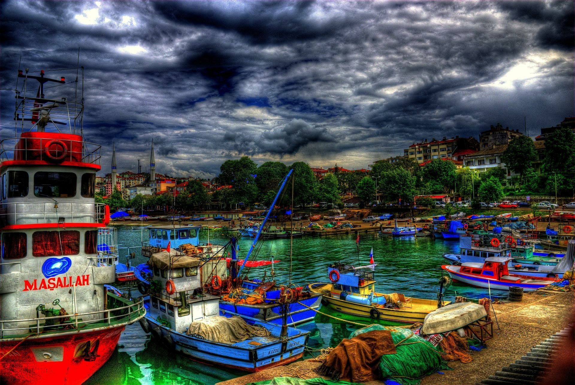 A vibrant marina in Turkey, showcasing colorful boats docked by lush trees under dramatic clouds. This HDR photography captures the serene yet lively atmosphere of the harbor.