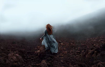 A redheaded woman is walking away through a foggy, rocky terrain. The scene, captured from the rear, creates a mystic and serene atmosphere. HD desktop wallpaper and background.