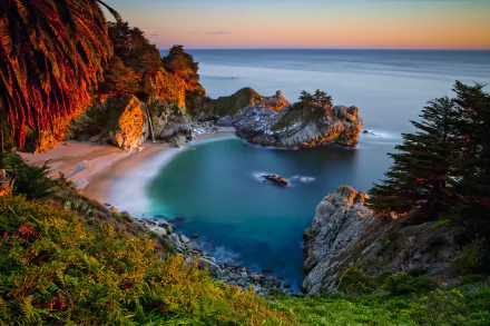 A serene beach scene in Big Sur, California, features a stunning coastline with a gentle waterfall cascading into the ocean, surrounded by lush greenery and tranquil waters.