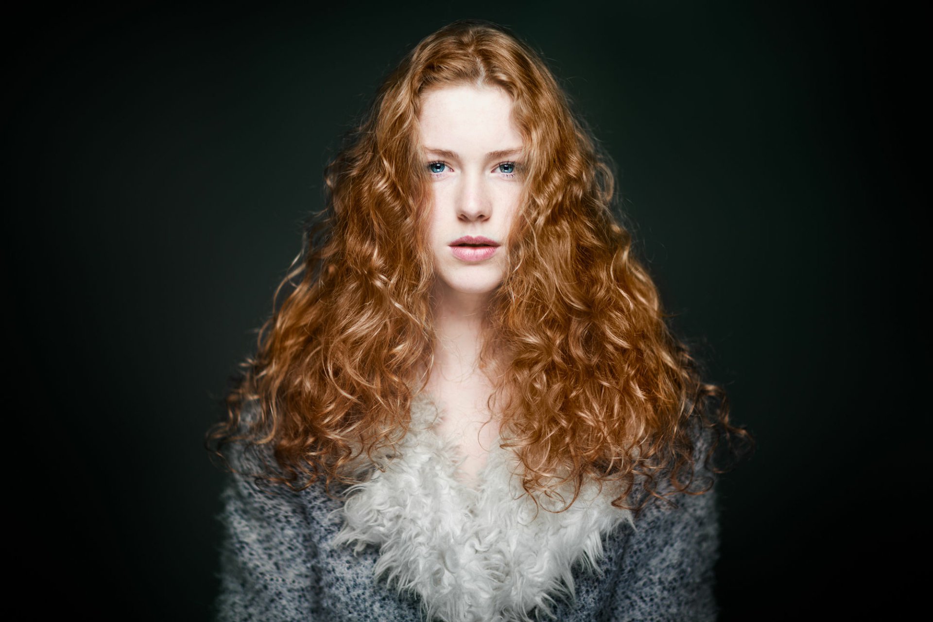 HD desktop wallpaper featuring a redhead woman model with striking blue eyes and voluminous curly hair against a dark background.