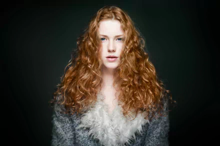HD desktop wallpaper featuring a redhead woman model with striking blue eyes and voluminous curly hair against a dark background.