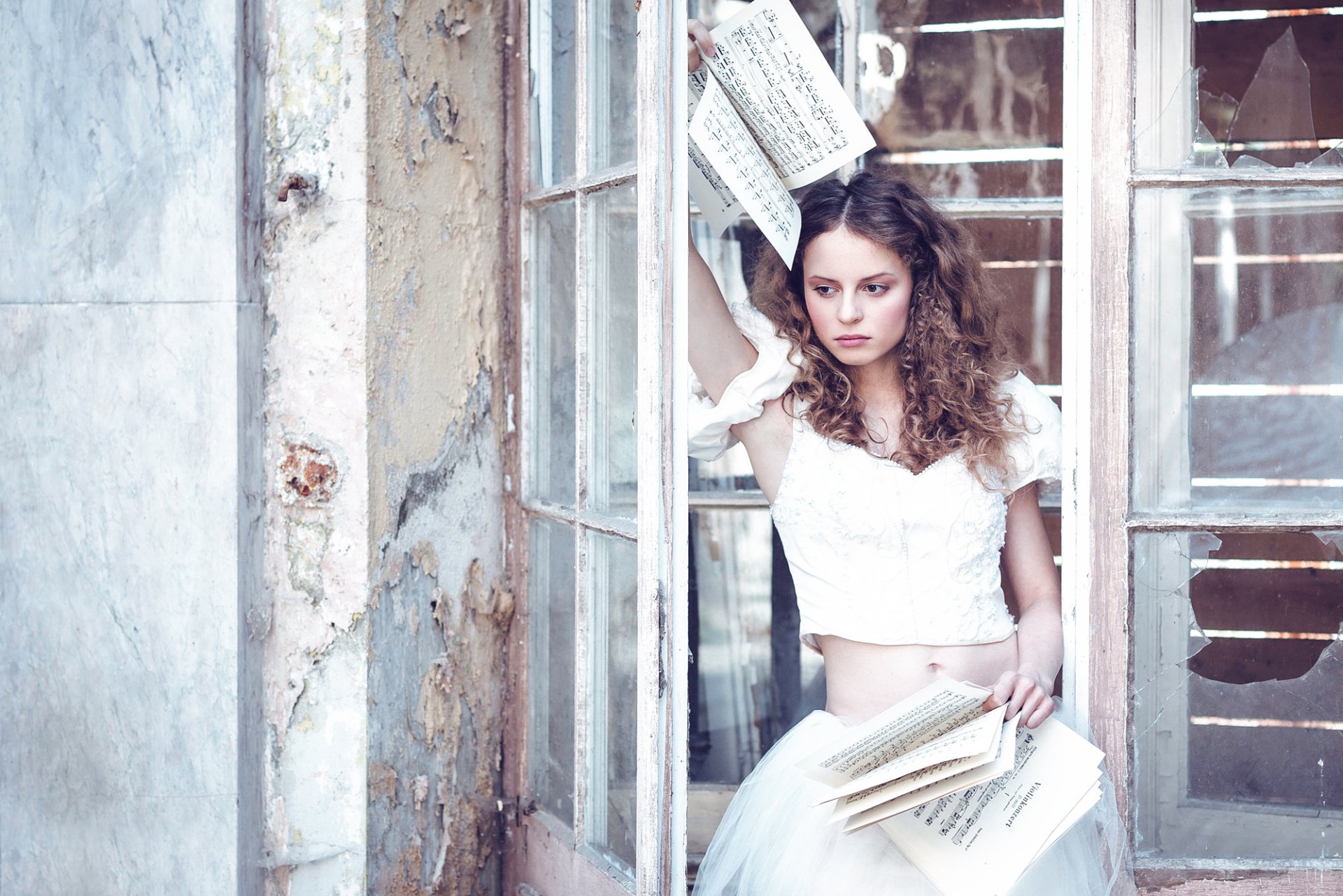 HD PC desktop wallpaper of a brunette woman model with brown eyes in a white dress, holding sheet music and surrounded by pages near a weathered window frame.