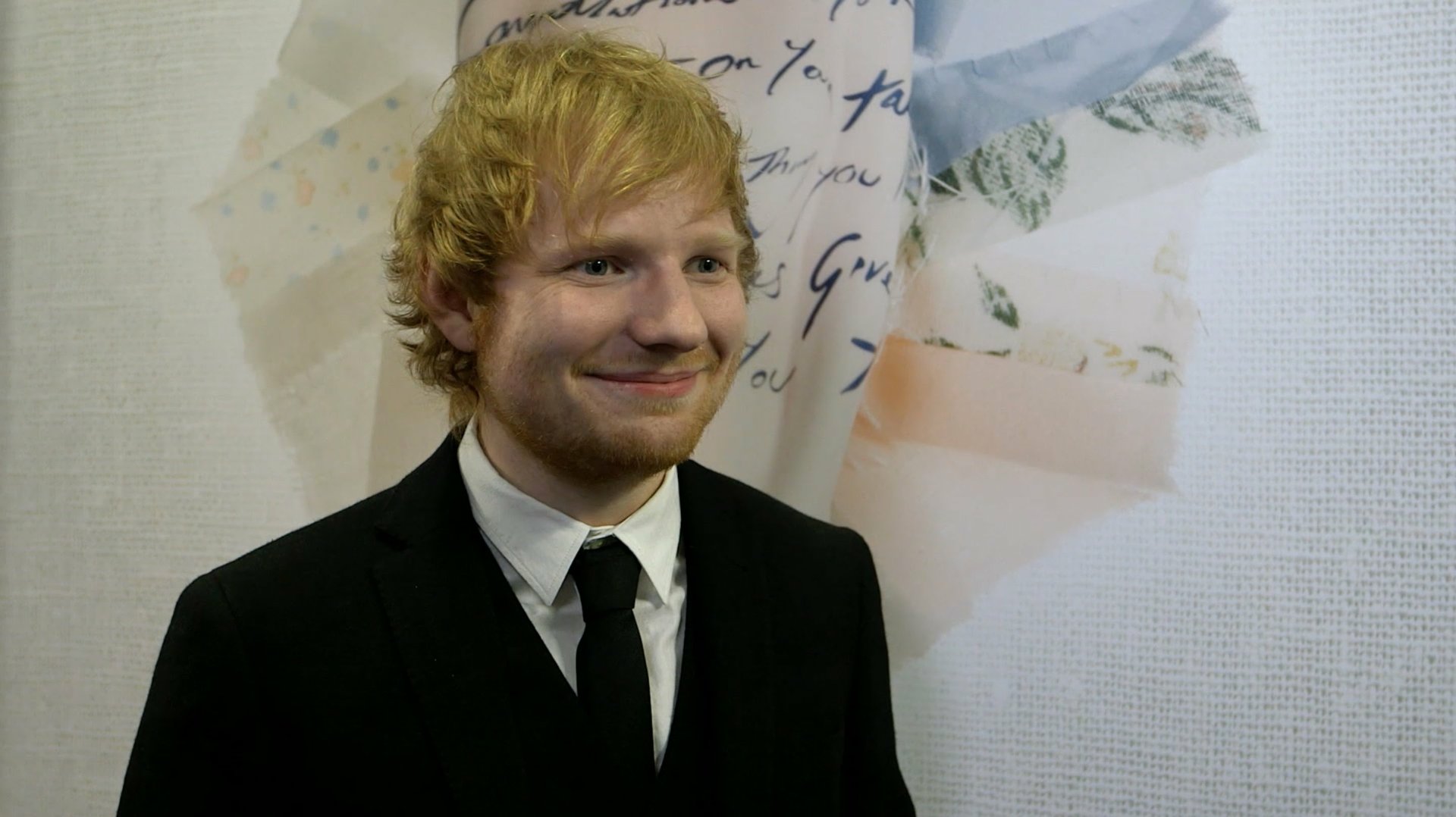 Ed Sheeran, the English singer, stands in front of a background with handwritten notes and flowers, wearing a black suit and tie. HD desktop wallpaper.