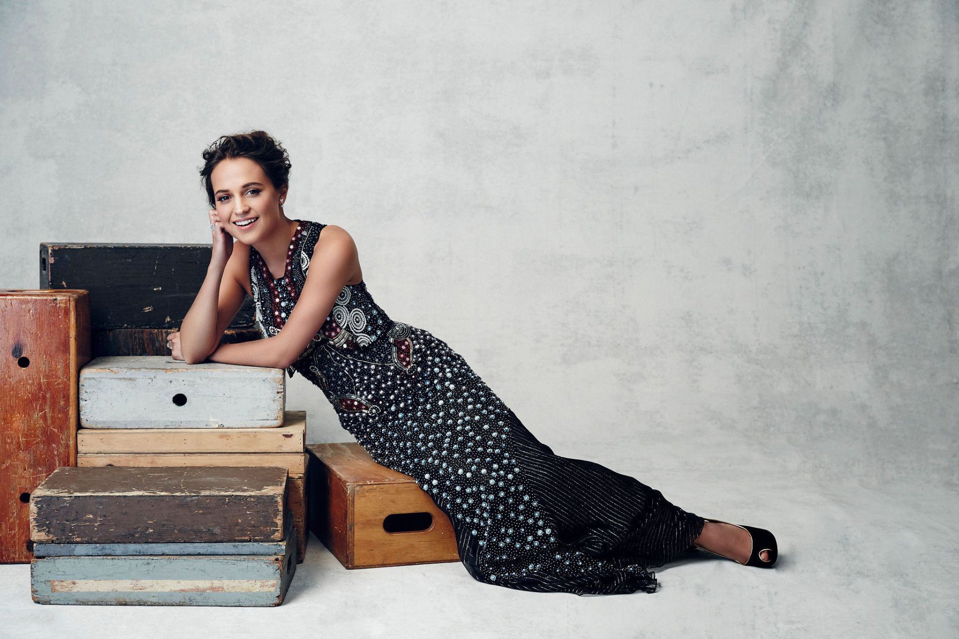Alicia Vikander, Swedish actress with brunette hair and brown eyes, smiles gracefully while leaning on stacked vintage suitcases against a neutral backdrop in this HD desktop wallpaper.