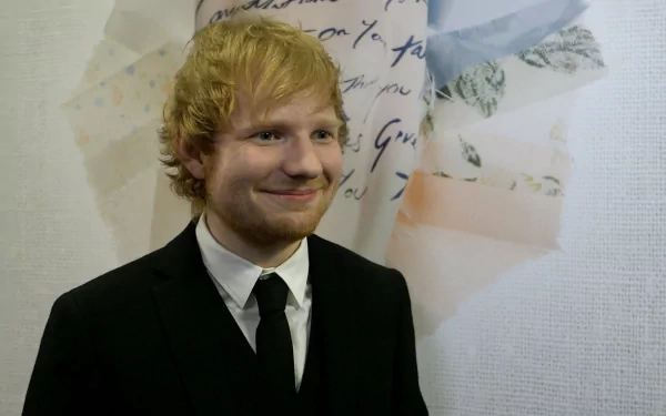 Ed Sheeran, the English singer, stands in front of a background with handwritten notes and flowers, wearing a black suit and tie. HD desktop wallpaper.