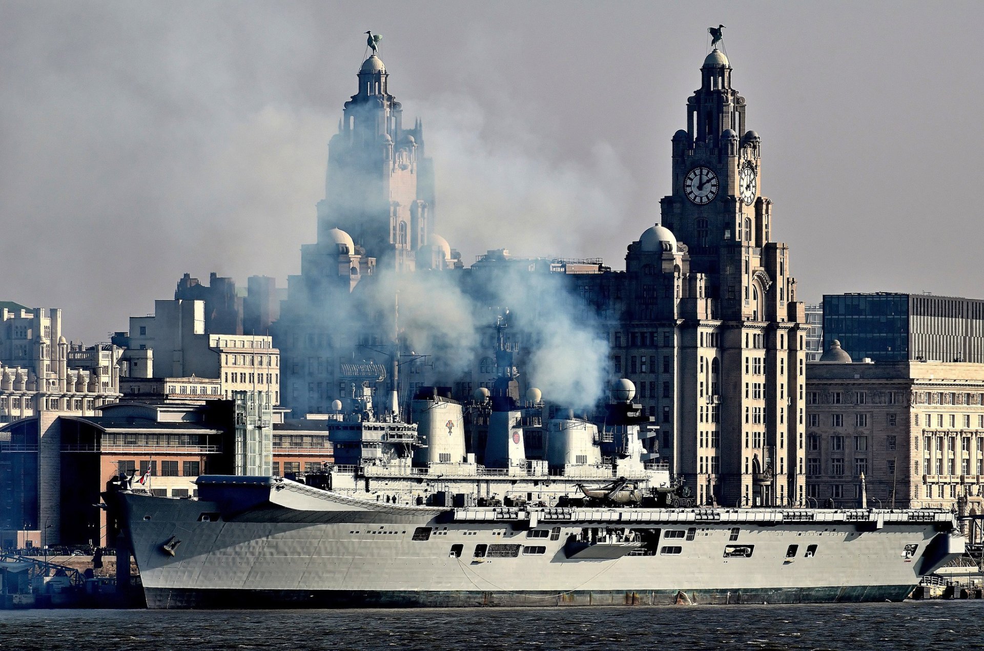 HD PC desktop wallpaper of Royal Navy aircraft carrier HMS Illustrious (R06) off a city waterfront, helicopters on deck and smoke rising.