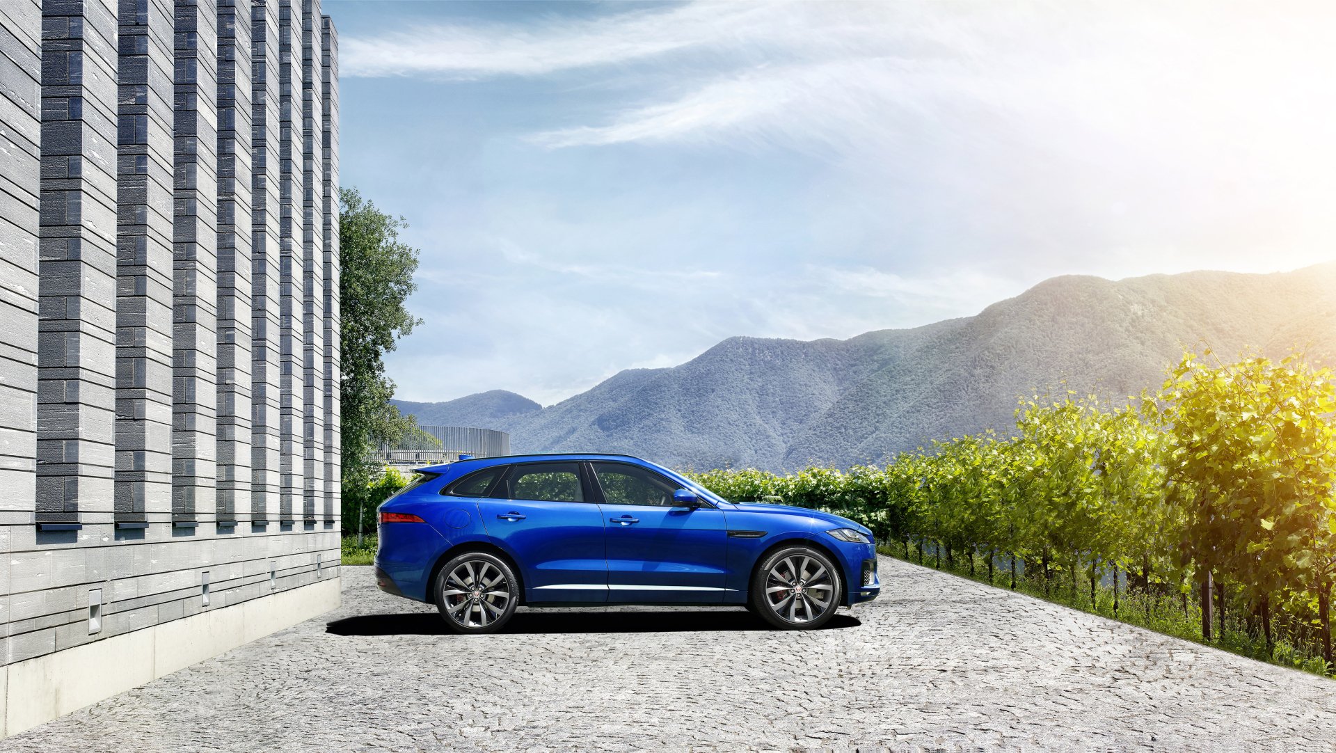A blue Jaguar F-PACE SUV parked on a paved area near a modern building, with mountains and greenery in the background, captured in 4K Ultra HD.