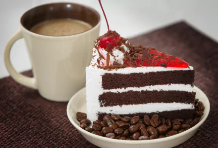 A slice of chocolate cake topped with cherry and whipped cream, surrounded by coffee beans, next to a steaming cup of coffee on a textured surface.