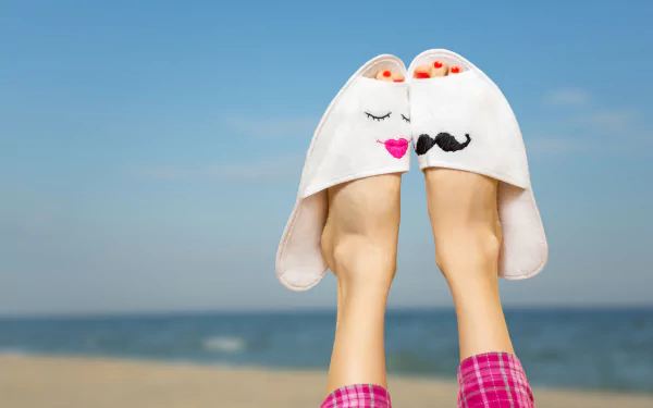 A woman’s feet adorned with playful slippers are raised on a sunlit beach, capturing the essence of summer and relaxation against a serene ocean background.