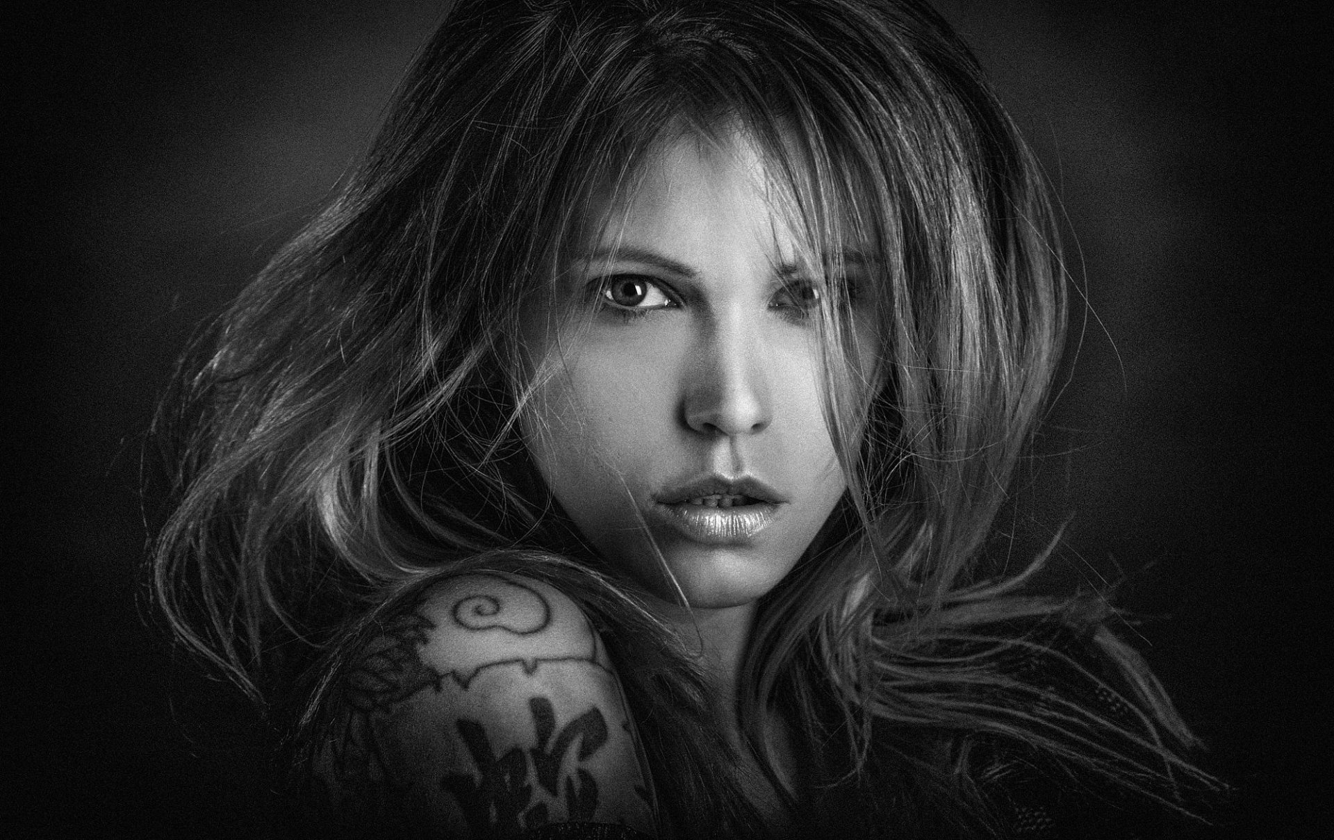 Black-and-white close-up of a tattooed female model with windblown hair and an intense gaze — HD PC desktop wallpaper background.